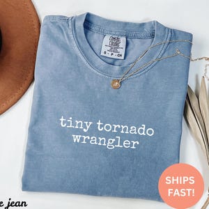 Puede incluir: Una camiseta azul claro con el texto "tiny tornado wrangler" impreso en blanco. La camiseta está hecha de una tela suave y cómoda y tiene un corte holgado.