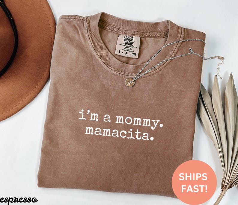Puede incluir: Camiseta color espresso con el texto "i'm a mommy. mamacita." Un collar plateado con un colgante descansa sobre la camiseta. La camiseta tiene una etiqueta Comfort Colors. Un sombrero marr&oacute;n y hojas secas est&aacute;n en el fondo.