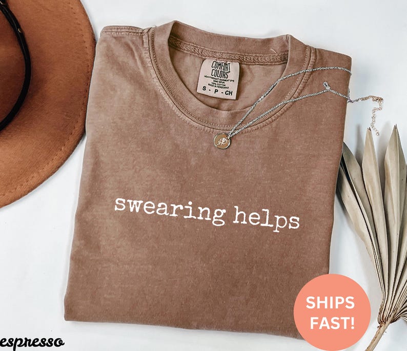May include: A brown t-shirt with the text "swearing helps" printed in white on the front. The shirt is made of a soft, comfortable material.