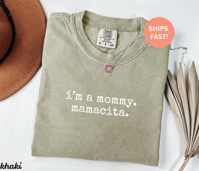 Puede incluir: Camiseta Comfort Colors color caqui con el texto "i'm a mommy. mamacita." impreso en blanco. La camiseta tiene cuello redondo y mangas cortas. Un collar plateado con un colgante est&aacute; sobre la camiseta.