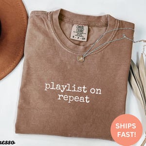 May include: A brown t-shirt with the text "playlist on repeat" in white lettering. The shirt is made of a soft, comfortable material and has a relaxed fit.