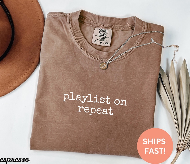 May include: A brown t-shirt with the text "playlist on repeat" in white lettering. The shirt is made of a soft, comfortable material and has a relaxed fit.