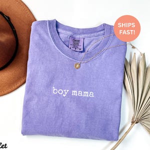 May include: A purple t-shirt with the text "boy mama" embroidered in white on the front. The shirt is made of a soft, comfortable material and has a relaxed fit.