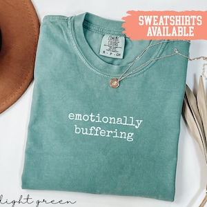 May include: Light green t-shirt with the phrase "emotionally buffering" in white text. A gold-coloured necklace with a pendant rests on the shirt. The shirt is labelled "Comfort Colors" and "sweatshirts available".