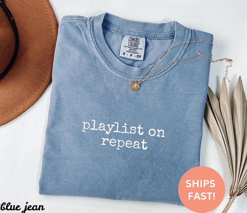 May include: A light blue t-shirt with the text "playlist on repeat" in white. The shirt is made of a soft, comfortable material.