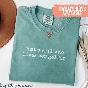 May include: Light green t-shirt with the text "just a girl who loves her golden" in white. A gold-coloured necklace with a pendant is draped across the shirt. The shirt has a Comfort Colors tag. The words "SWEATSHIRTS AVAILABLE" are in a peach-coloured box.
