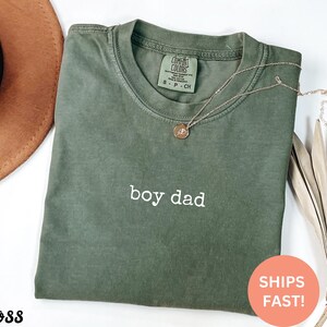 May include: A green t-shirt with the text "boy dad" in white lettering. The shirt is made of a soft, comfortable material and has a relaxed fit.