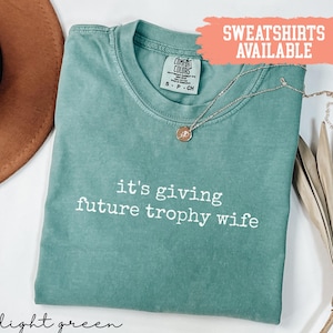 May include: Light green t-shirt with the phrase "it's giving future trophy wife" in white text. A gold necklace with a pendant rests on the shirt. The shirt has a Comfort Colors tag.