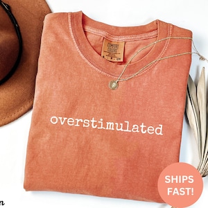 K&ouml;nnte beinhalten: Ein korallenfarbenes T-Shirt mit dem Aufdruck "overstimulated" in Wei&szlig;. Das Shirt ist aus weichem, bequemem Stoff und hat eine lockere Passform.