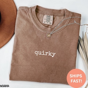 May include: Espresso-colored t-shirt with the word "quirky" in white. A silver necklace with a pendant rests on the shirt. The shirt has a Comfort Colors tag. A brown hat and dried plant are in the background.
