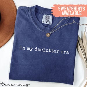 May include: A navy blue t-shirt with the text "in my declutter era" in white. The shirt has a crew neck and short sleeves. A gold necklace with a pendant is draped across the shirt. A peach banner reads "Sweatshirts Available".