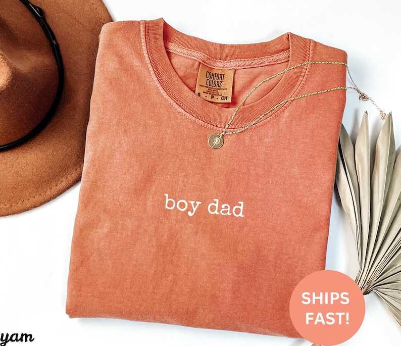 May include: A coral-colored t-shirt with the text "boy dad" embroidered in white. The shirt is made of a soft, comfortable fabric and has a relaxed fit.