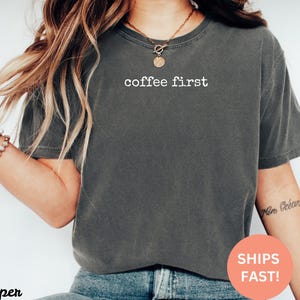 May include: Dark gray t-shirt with the text "coffee first" in white. The shirt is a crew neck style and appears to be made of a soft, comfortable material. The shirt is worn with blue jeans.
