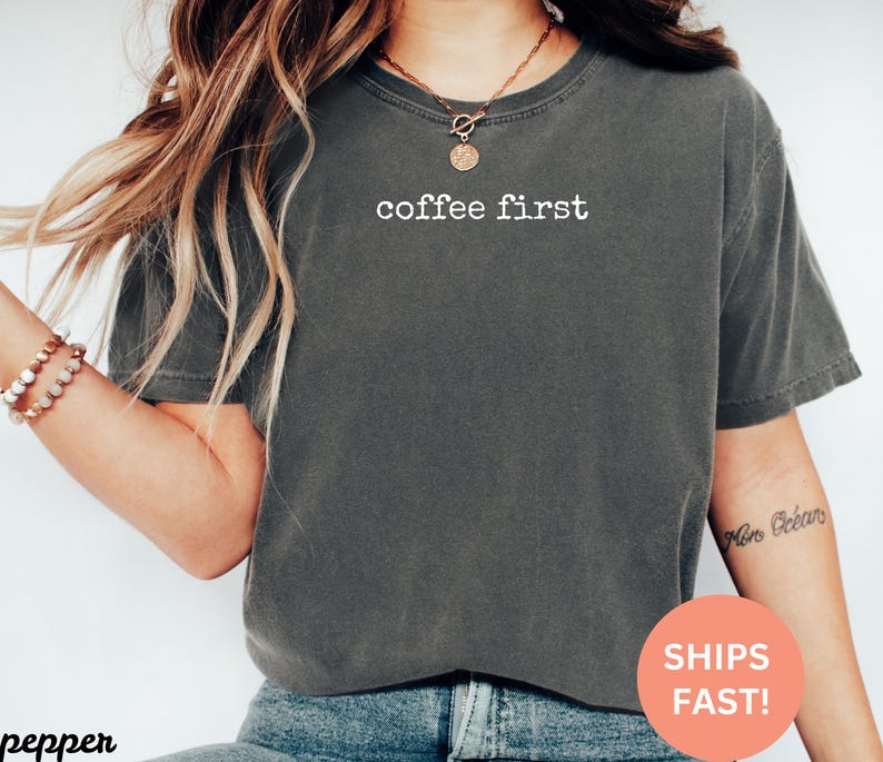 May include: Dark gray t-shirt with the text "coffee first" in white. The shirt is a crew neck style and appears to be made of a soft, comfortable material. The shirt is worn with blue jeans.