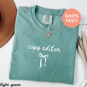 Peut inclure: T-shirt vert clair avec l'inscription "copy editor" en écriture blanche et un motif de nœud. Le t-shirt a un col rond et des manches courtes. Un collier argenté avec un pendentif est posé sur le t-shirt.