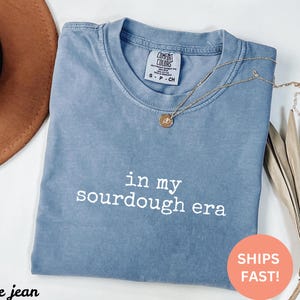 May include: A blue jean t-shirt with the text "in my sourdough era" printed in white on the front.