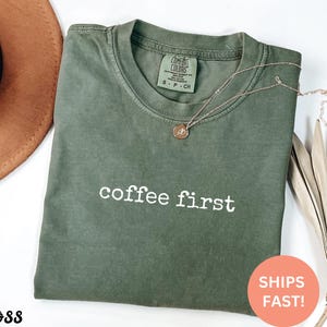 May include: Olive green t-shirt with the phrase "coffee first" in white block letters. A gold necklace with a small pendant rests on the shirt. The shirt is folded, and a brown hat is visible in the background.