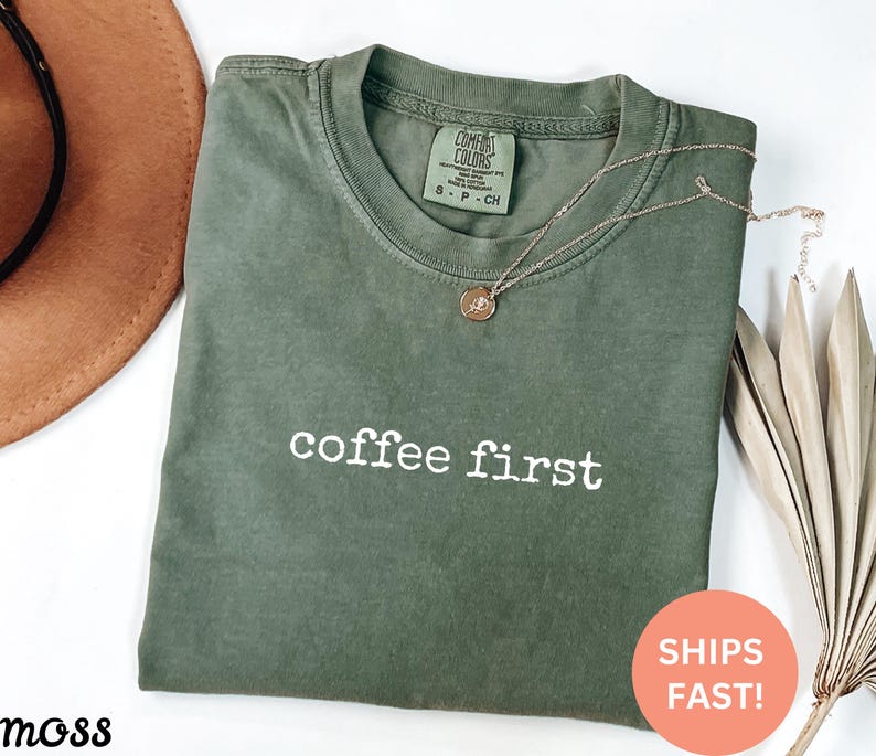 May include: Olive green t-shirt with the phrase "coffee first" in white block letters. A gold necklace with a small pendant rests on the shirt. The shirt is folded, and a brown hat is visible in the background.