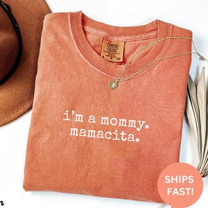 Puede incluir: Una camiseta Comfort Colors de color coral con la frase "i'm a mommy. mamacita." en letras blancas. Un collar dorado con un colgante descansa sobre la camiseta. Un sombrero marr&oacute;n es visible en el fondo.