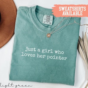 Può includere: T-shirt verde chiaro con la scritta bianca "just a girl who loves her pointer". Una collana d'oro con un ciondolo è drappeggiata sulla maglietta. La maglietta è etichettata "Comfort Colors" e "Sweatshirts Available".
