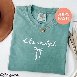 May include: Light green t-shirt featuring the words "data analyst" in white script and a bow graphic. The shirt has a crew neck and short sleeves. A gold necklace with a pendant is draped on the shirt.