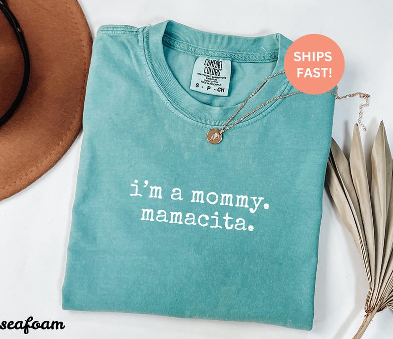 Puede incluir: Camiseta verde mar con el texto "i'm a mommy. mamacita." Un collar dorado con un colgante est&aacute; sobre la camiseta. La camiseta tiene una etiqueta Comfort Colors. Un sombrero marr&oacute;n y hojas secas est&aacute;n en el fondo.