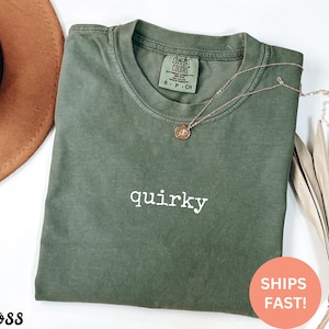 May include: An olive green t-shirt with the word "quirky" in white script. A gold necklace with a pendant rests on the shirt. The shirt is folded and has a tag that says "Comfort Colors". A round sticker says "Ships Fast!"