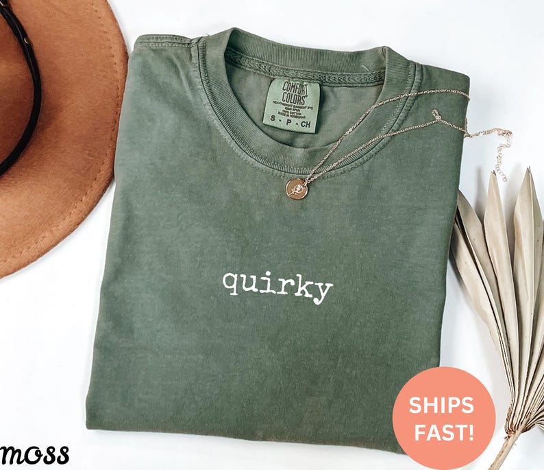 May include: An olive green t-shirt with the word "quirky" in white script. A gold necklace with a pendant rests on the shirt. The shirt is folded and has a tag that says "Comfort Colors". A round sticker says "Ships Fast!"