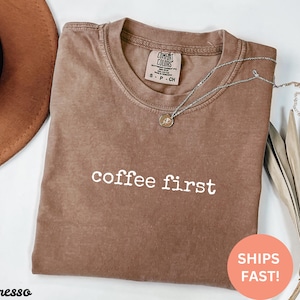 May include: Espresso-colored t-shirt with the phrase "coffee first" in white lettering. A silver necklace with a small pendant rests on the shirt. The shirt is folded, and a brown hat is visible in the background.