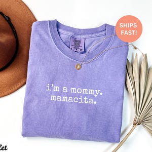 Puede incluir: Una camiseta violeta Comfort Colors con el texto "i'm a mommy. mamacita." en blanco. Un collar dorado con un colgante est&aacute; sobre la camiseta. Un sombrero marr&oacute;n y hojas de palma secas est&aacute;n en el fondo.