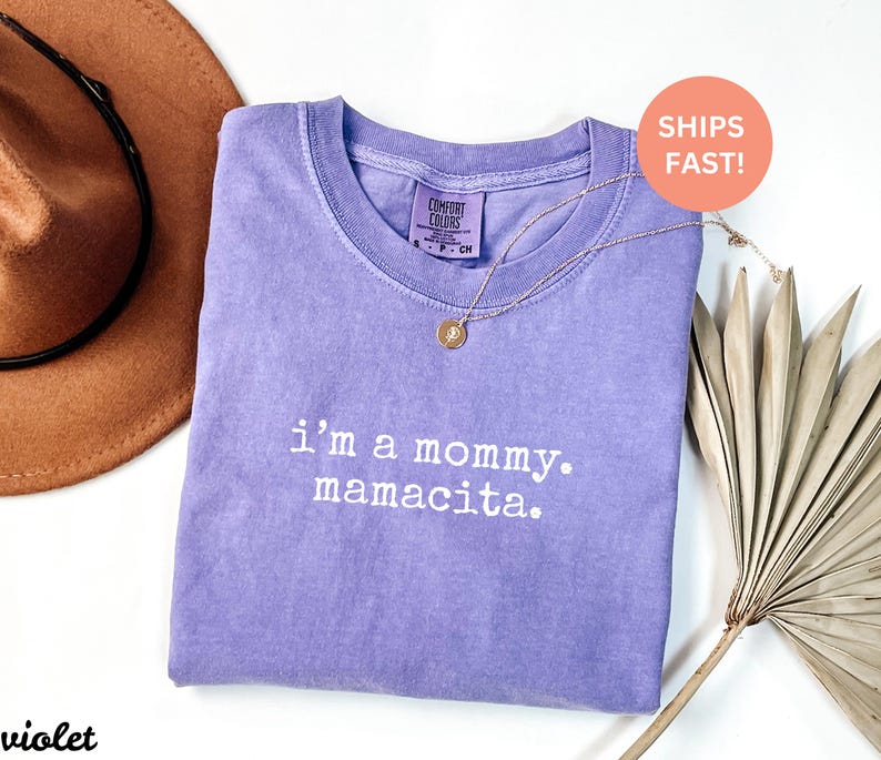 Puede incluir: Una camiseta violeta Comfort Colors con el texto "i'm a mommy. mamacita." en blanco. Un collar dorado con un colgante est&aacute; sobre la camiseta. Un sombrero marr&oacute;n y hojas de palma secas est&aacute;n en el fondo.