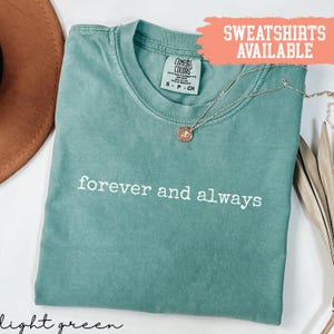 Può includere: T-shirt verde chiaro con le parole "forever and always" in bianco. Una collana color oro con un ciondolo poggia sulla maglietta. La maglietta è del marchio Comfort Colors e il testo "SWEATSHIRTS AVAILABLE" è in un riquadro color pesca.