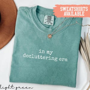 Op de afbeelding: Lichtgroen Comfort Colors T-shirt met de witte tekst "in my decluttering era". Een gouden ketting met een hanger ligt op het shirt. De woorden "Sweatshirts Available" staan in een perzikkleurige doos.