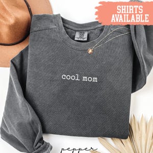 May include: A charcoal gray sweatshirt with the embroidered text "cool mom" in white. A gold necklace with a pendant is draped on the shirt. The words "SHIRTS AVAILABLE" are in a coral-colored banner.