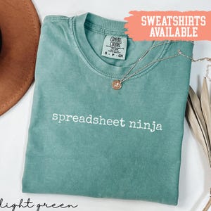 May include: Light green t-shirt with the words "spreadsheet ninja" in white. A silver necklace with a pendant rests on the shirt. The shirt is labeled "Comfort Colors" and "sweatshirts available" is written in a peach-colored box.