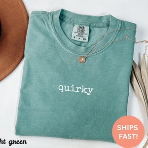 May include: A light green t-shirt with the word "quirky" in white script. A gold necklace with a pendant is draped over the shirt. The shirt is folded and placed on a white surface.