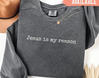 May include: A dark grey sweatshirt with the white text "Jesus is my reason". The crew neck sweatshirt has long sleeves. A gold necklace with a pendant is draped on the shirt. The words "Shirts Available" are in a red banner.