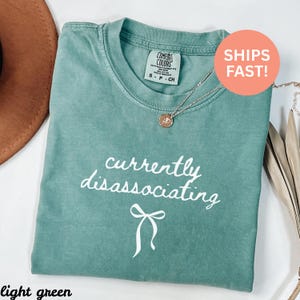 Könnte beinhalten: Hellgrünes T-Shirt mit dem Schriftzug "currently disassociating" in weißer Schrift und einer Schleifengrafik. Eine goldfarbene Halskette mit Anhänger ist auf dem T-Shirt drapiert. Das T-Shirt ist mit "Comfort Colors" gekennzeichnet.