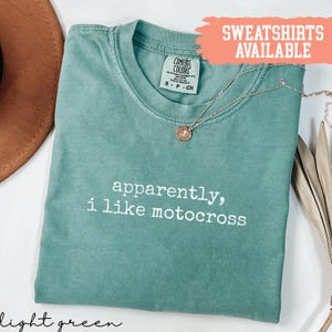 Könnte beinhalten: Hellgrünes T-Shirt mit dem weißen Text "apparently, i like motocross". Eine goldene Halskette mit Anhänger liegt auf dem Shirt. Das Shirt ist mit "Comfort Colors" und "S-P-CH" gekennzeichnet.