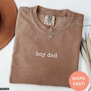 May include: A brown t-shirt with the text "boy dad" in white lettering. The shirt is made of a soft, comfortable material and has a relaxed fit.