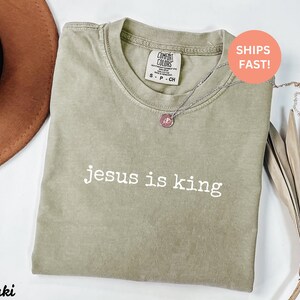 K&ouml;nnte beinhalten: Ein khakifarbenes T-Shirt mit dem Aufdruck "jesus is king" in Wei&szlig; auf der Vorderseite.