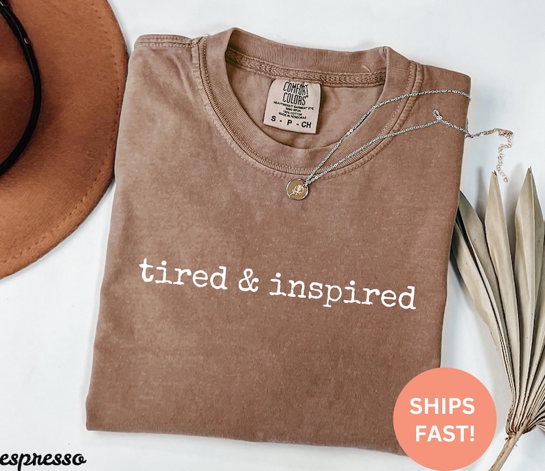 May include: A brown t-shirt with the text "tired & inspired" printed in white. The shirt is made of a soft, comfortable material.