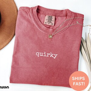 May include: Crimson-colored t-shirt with the word "quirky" in white. The shirt has a crew neck and a small tag at the neckline. A gold necklace with a pendant is draped across the shirt. A brown hat is partially visible in the upper left corner.