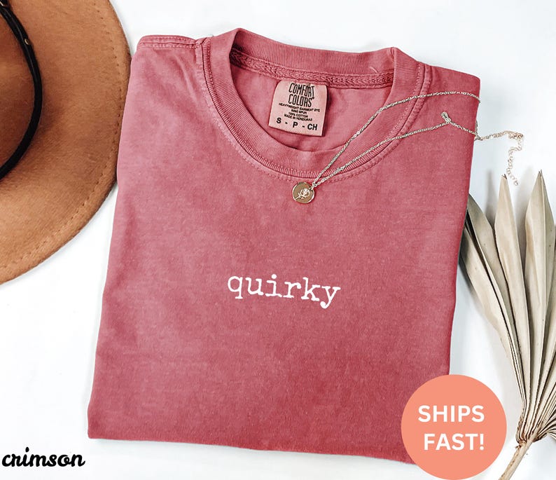 May include: Crimson-colored t-shirt with the word "quirky" in white. The shirt has a crew neck and a small tag at the neckline. A gold necklace with a pendant is draped across the shirt. A brown hat is partially visible in the upper left corner.