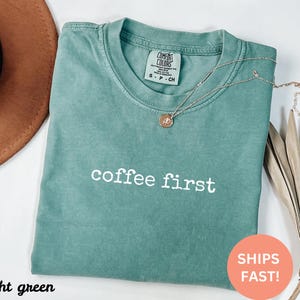 May include: Light green t-shirt with the text "coffee first" in white. The shirt has a crew neck and short sleeves. A gold necklace with a pendant is draped on the shirt. A brown hat is in the upper left corner.