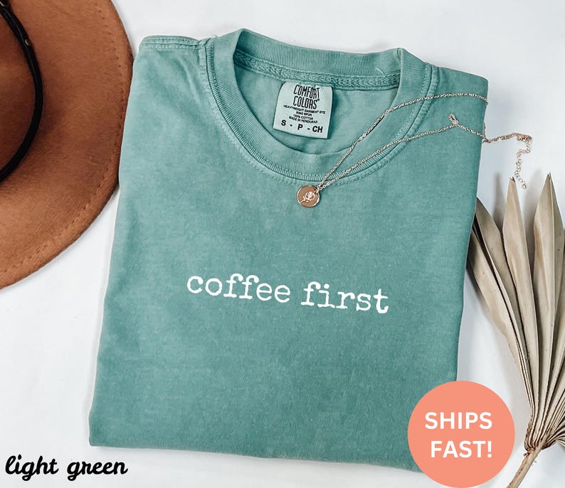 May include: Light green t-shirt with the text "coffee first" in white. The shirt has a crew neck and short sleeves. A gold necklace with a pendant is draped on the shirt. A brown hat is in the upper left corner.