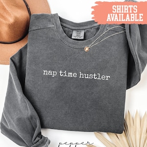 Pode incluir: Uma camisola cinza escuro com a frase "nap time hustler" em branco. A camisola tem gola redonda e mangas compridas. Um colar de ouro com um pingente está sobre a camisa. Uma faixa vermelha diz "Shirts Available".