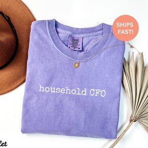 Może przedstawiać: Fioletowy T-shirt z nadrukiem "household CFO" w kolorze białym. Koszulka wykonana jest z miękkiego, wygodnego materiału.