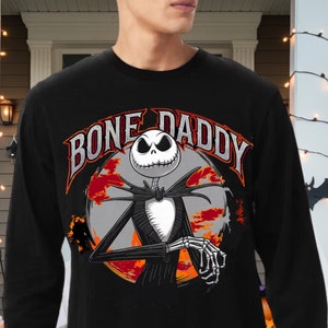 Może przedstawiać: Czarny T-shirt z długim rękawem z grafiką Jacka Skellingtona z filmu Koszmar przed świętami. Grafika przedstawia Jacka Skellingtona z tekstem "Bone Daddy" wok&oacute;ł niego.