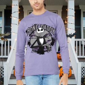 Może przedstawiać: Fioletowy T-shirt z długim rękawem z grafiką Jacka Skellingtona z filmu Koszmar przed świętami. Grafika zawiera tekst "Bone Daddy".